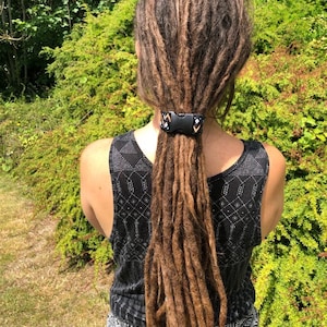 K&ouml;nnte beinhalten: Eine Person mit langen braunen Dreadlocks, die mit einem schwarzen und wei&szlig;en gemusterten Haargummi zur&uuml;ckgebunden sind. Das Haargummi hat eine schwarze Schnalle.