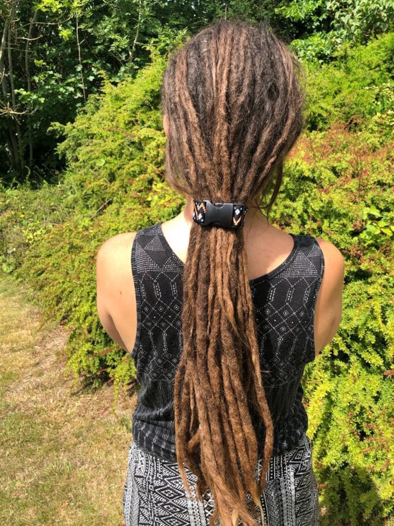 K&ouml;nnte beinhalten: Eine Person mit langen braunen Dreadlocks, die mit einem schwarzen und wei&szlig;en gemusterten Haargummi zur&uuml;ckgebunden sind. Das Haargummi hat eine schwarze Schnalle.