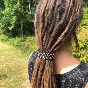 K&ouml;nnte beinhalten: Ein schwarzes Perlenhaarband mit einem geometrischen Muster in Orange, Wei&szlig; und Blau. Das Haarband ist um einen Pferdeschwanz aus langen braunen Dreadlocks gebunden.