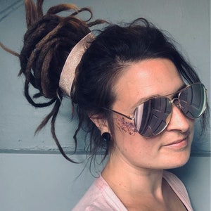 Könnte beinhalten: Eine Frau mit braunen Dreadlocks, die zu einem Dutt mit einem silbernen Stirnband gebunden sind. Sie trägt eine verspiegelte Aviator-Sonnenbrille und hat Glitzer auf der Wange.