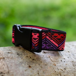 K&ouml;nnte beinhalten: Eine schwarze verstellbare Schnalle mit einem bunten gewebten Band. Das Band hat ein geometrisches Muster in den Farben Pink, Rot, Lila und Schwarz.