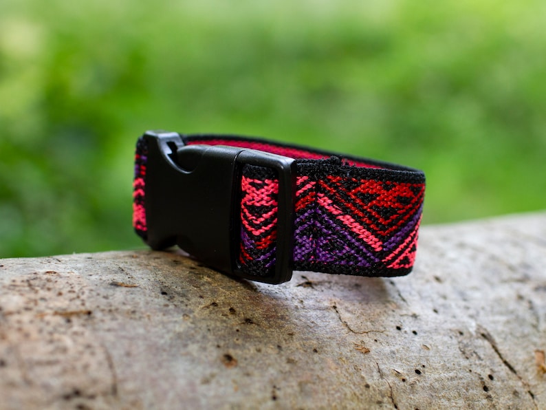 K&ouml;nnte beinhalten: Eine schwarze verstellbare Schnalle mit einem bunten gewebten Band. Das Band hat ein geometrisches Muster in den Farben Pink, Rot, Lila und Schwarz.