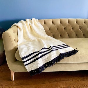 May include: A white crocheted throw blanket with black stripes and fringe. The blanket is draped over a light brown couch.