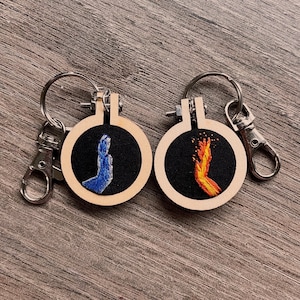 Op de afbeelding: Twee houten sleutelhangers met een borduurring en een zwarte achtergrond. De ene sleutelhanger heeft een blauwe vlam en de andere een oranje vlam.