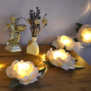 Op de afbeelding: Een string met sprookjeslichtjes met witte papieren bloemen, elk met een warm wit LED-lichtje erin. De lichtjes zijn gespannen over een houten oppervlak met een gouden cherubbeeld en een vaas met gedroogde lavendel op de achtergrond.