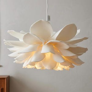 May include: A white pendant light fitting shaped like a blooming flower with multiple overlapping petals. The lamp is suspended by a white cord and emits a warm, soft glow from its centre, illuminating the layered structure of the petals.