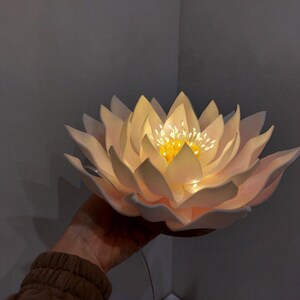 May include: A white paper flower with a yellow center and a string of lights inside. The flower is lit up and looks like a lotus flower.