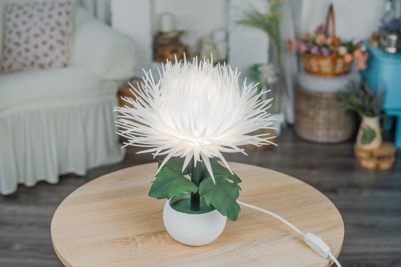 May include: A white paper flower lamp with green leaves in a white ceramic pot. The lamp is on a wooden table.