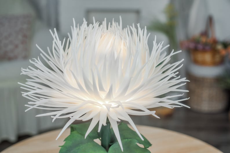 May include: A white paper flower with a light inside, resembling a chrysanthemum. The flower is on a table with green leaves.