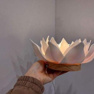 May include: A white ceramic lotus flower lamp with a wooden base. The lamp is lit and casts a warm glow.