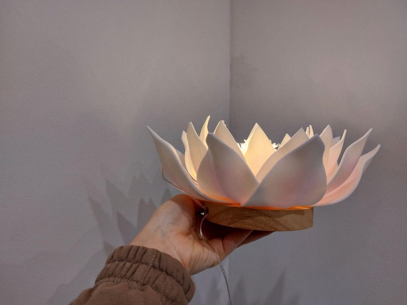 May include: A white ceramic lotus flower lamp with a wooden base. The lamp is lit and casts a warm glow.