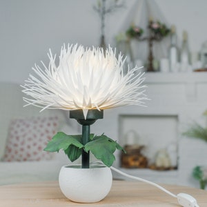 May include: White paper flower lamp with green leaves in a white pot. The lamp is on a wooden table and has a white cord.
