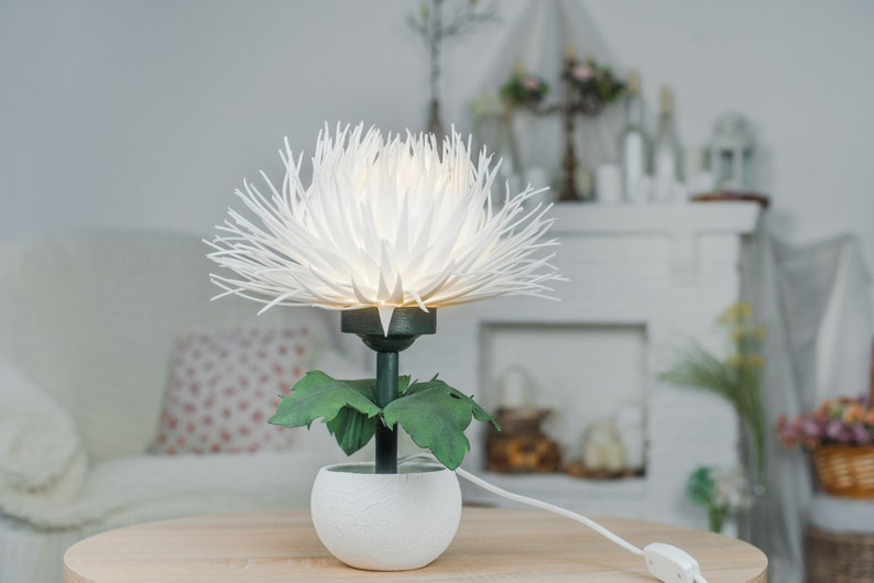 May include: White paper flower lamp with green leaves in a white pot. The lamp is on a wooden table and has a white cord.
