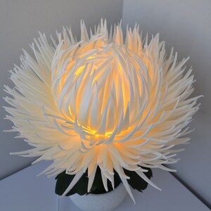 May include: A white, 3D printed flower lamp with a warm, yellow light emanating from the center. The flower is made of many thin, delicate petals that create a soft, airy look.