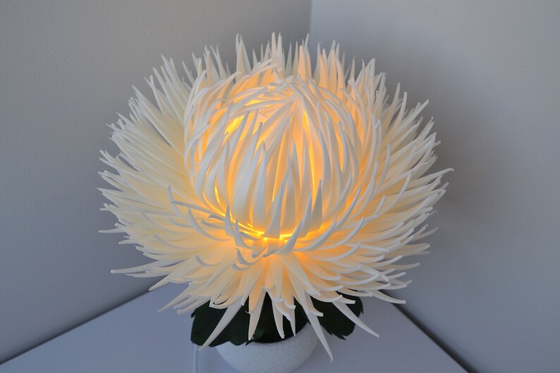 May include: A white, 3D printed flower lamp with a warm, yellow light emanating from the center. The flower is made of many thin, delicate petals that create a soft, airy look.