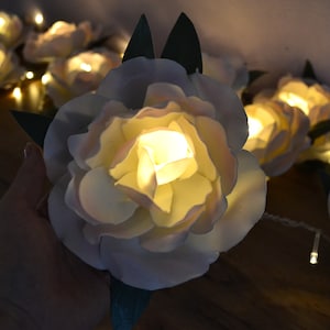Op de afbeelding: Een string met witte bloemen van stof met warm witte LED-lampjes. De bloemen zijn op een draad met groene bladeren geregen.