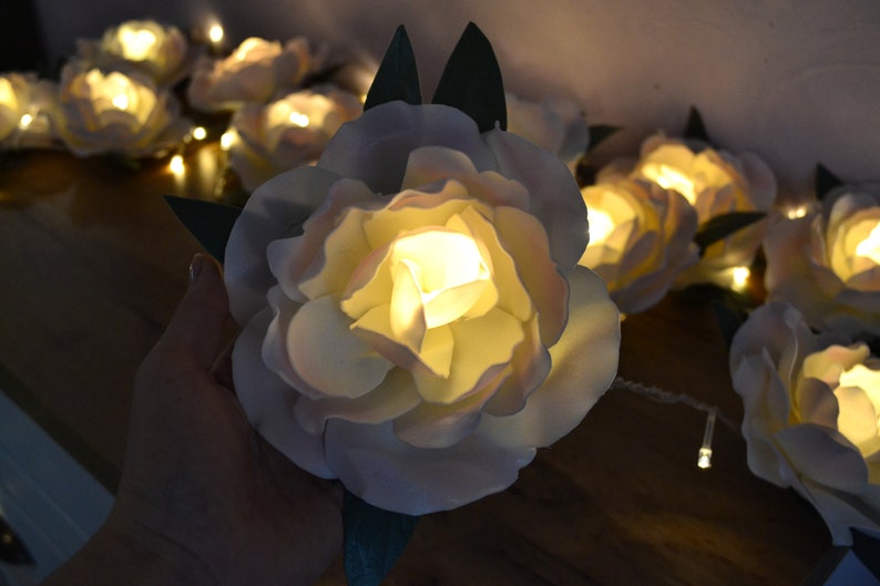 Op de afbeelding: Een string met witte bloemen van stof met warm witte LED-lampjes. De bloemen zijn op een draad met groene bladeren geregen.