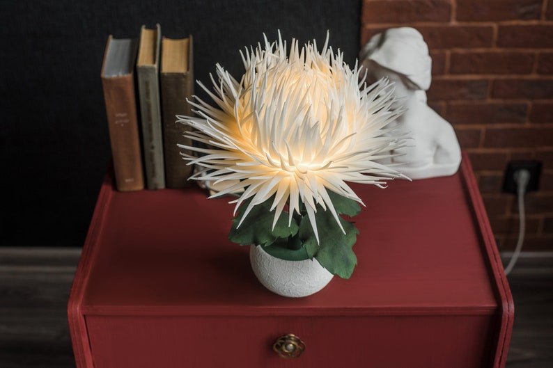 May include: A white paper flower lamp with green leaves in a white pot on a red nightstand. The lamp is lit and casting a warm glow.