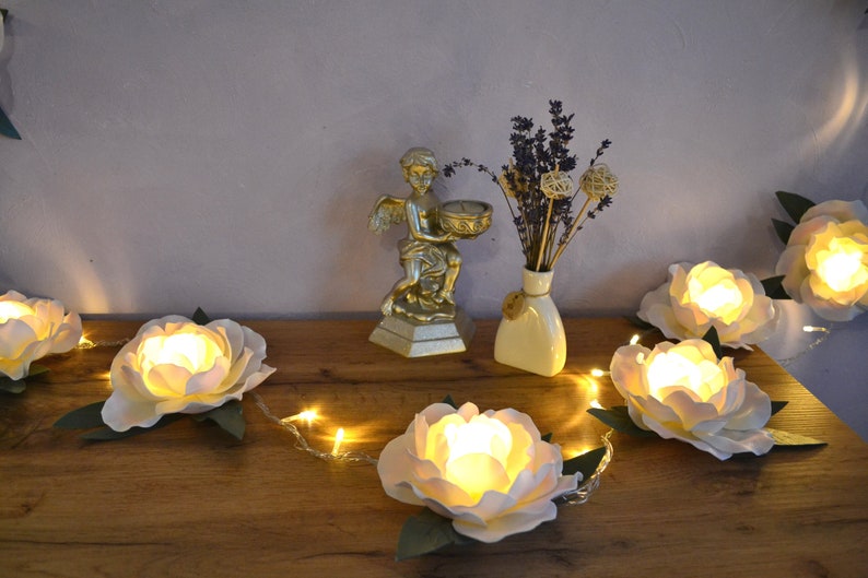 Op de afbeelding: Een string met sprookjeslichtjes met witte papieren bloemen, elk met een warm wit LED-lichtje erin. De lichtjes zijn gespannen over een houten oppervlak met een gouden cherubbeeld en een vaas met gedroogde lavendel op de achtergrond.