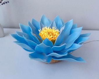 以下が含まれることがあります： 黄色い中心部を持つ、水色の蓮の花の形をしたランプ。 ランプは柔らかい素材でできており、電源用のワイヤーが付いています。 ランプは白い表面にあります。