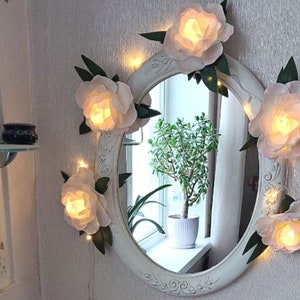 Puede incluir: Un espejo ovalado blanco con un marco decorativo adornado con flores de papel blanco y luces de cadena. El espejo refleja una planta en maceta y una ventana.