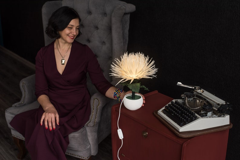 May include: A woman in a burgundy dress sits in a gray armchair, looking at a white paper flower lamp on a red table. The lamp is lit and casts a warm glow. A vintage typewriter sits on the table.