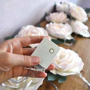 Op de afbeelding: Een wit batterijpakket met een knop aan de zijkant, gebruikt om een ​​lichtsnoer van stroom te voorzien. De lampjes zijn bevestigd aan kunstbloemen, die wit zijn met een vleugje roze.