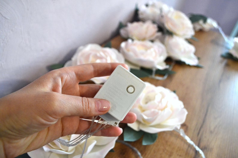 Op de afbeelding: Een wit batterijpakket met een knop aan de zijkant, gebruikt om een ​​lichtsnoer van stroom te voorzien. De lampjes zijn bevestigd aan kunstbloemen, die wit zijn met een vleugje roze.