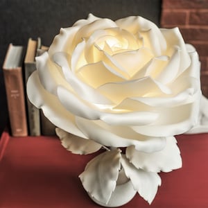 May include: A white paper rose lamp with a warm white light. The lamp is on a red table with books behind it.