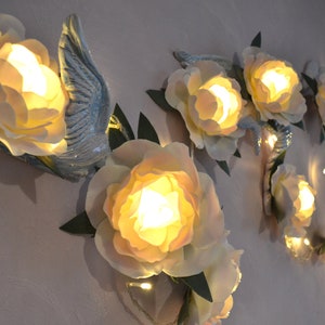 Op de afbeelding: Een wanddecoratie met een guirlande van witte papieren bloemen met lampjes erin. De bloemen zijn bevestigd aan een muur met groene bladeren en een blauwe vogel.