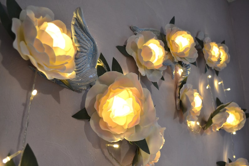 Op de afbeelding: Een wanddecoratie met een guirlande van witte papieren bloemen met lampjes erin. De bloemen zijn bevestigd aan een muur met groene bladeren en een blauwe vogel.