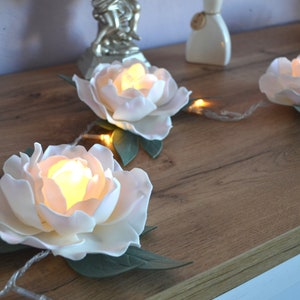 May include: A string of white faux flower lights with warm white LED bulbs. The flowers are arranged on a wooden surface.