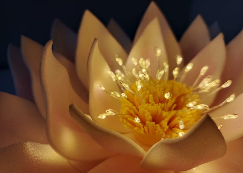 May include: A close-up of a white and yellow artificial lotus flower with a center of yellow stamens and white petals. The flower is illuminated with small white lights.