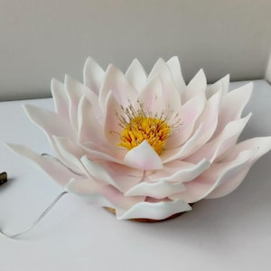 May include: A white and pink paper flower with a yellow center. The flower is lit up with a white cord and USB plug.