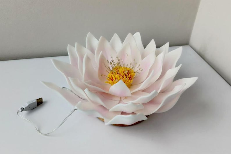 May include: A white and pink paper flower with a yellow center. The flower is lit up with a white cord and USB plug.