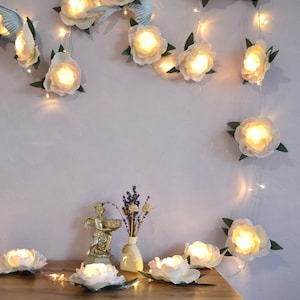 Op de afbeelding: Een lichtsnoer met witte papieren bloemen en warm witte LED-lampjes. De lichten zijn om een muur en op een houten tafel gehangen. De bloemen zijn in een cascade-achtige opstelling gerangschikt.
