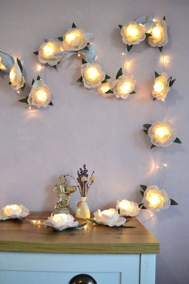 Op de afbeelding: Een lichtsnoer met witte papieren bloemen en warm witte LED-lampjes. De lichten zijn om een muur en op een houten tafel gehangen. De bloemen zijn in een cascade-achtige opstelling gerangschikt.