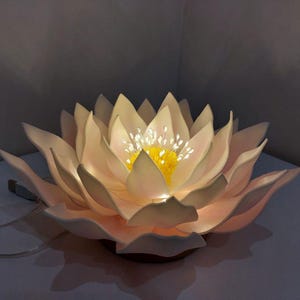 May include: A white, paper-like lotus flower lamp with a yellow center and white lights. The lamp is lit and casting a warm glow.
