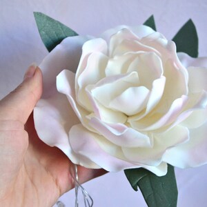 Op de afbeelding: Een witte en roze suikerbloem met groene bladeren. De bloem is gemaakt van fondant en is een populaire decoratie voor taarten en andere desserts.