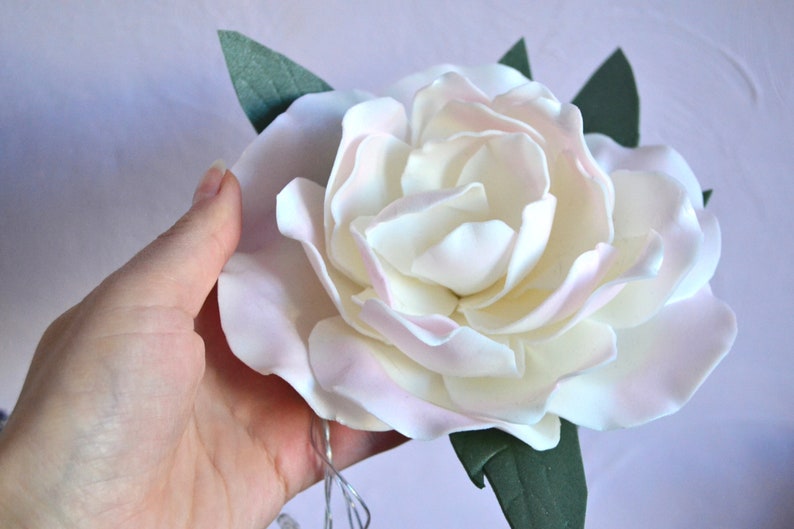 Op de afbeelding: Een witte en roze suikerbloem met groene bladeren. De bloem is gemaakt van fondant en is een populaire decoratie voor taarten en andere desserts.