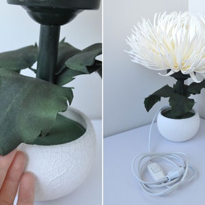 May include: A white table lamp with a white ceramic base and a white paper flower resembling a chrysanthemum. The lamp has green leaves and a black cord with a white switch.
