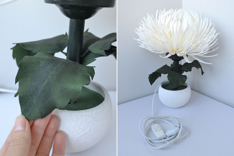 May include: A white table lamp with a white ceramic base and a white paper flower resembling a chrysanthemum. The lamp has green leaves and a black cord with a white switch.