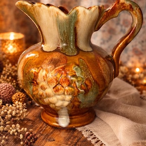 May include: A decorative ceramic pitcher with a bulbous body and a scalloped rim. The pitcher features a brown, green, and cream color scheme with a textured surface and a handle. The pitcher is likely used for serving beverages.