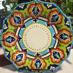 May include: A decorative ceramic plate with a scalloped edge. The plate features a vibrant design with a white center, a light blue ring, and a border of green, blue, red, and yellow floral patterns. The plate is displayed on a black metal stand.