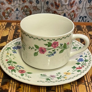 May include: A white teacup and saucer set with a floral design. The cup and saucer are decorated with pink, blue, and yellow flowers, and green leaves. The cup has a handle and sits on the matching saucer.