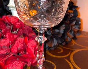 May include: A clear glass stemmed wine glass with a cut crystal design and a frosted flower etched on the bowl. The glass is sitting on a brown surface with gold swirls.