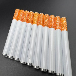 May include: A set of 10 white metal cigarette-shaped pipes with orange tips. The pipes are arranged in a row, with the tips facing the viewer.