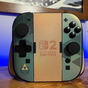 May include: A Nintendo Switch game console controller set featuring a custom skin with a green geometric pattern, black buttons, and a central gold panel displaying the Nintendo Switch logo and the number 2. A Triforce emblem is visible on the left.