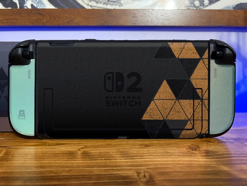 May include: A black Nintendo Switch console featuring a custom skin with a geometric triangle pattern in black and tan on the right side. The left and right edges are light mint green, and the center displays the Nintendo Switch 2 logo in black.