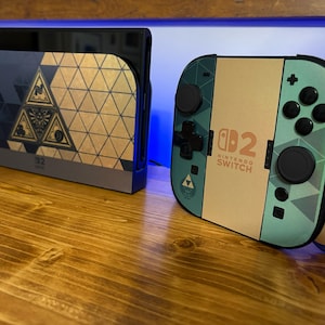 May include: A Nintendo Switch console dock and a Joy-Con controller grip featuring a custom skin with a geometric pattern in shades of teal, tan, and dark blue. The design includes a central triangular emblem and the text Nintendo Switch 2.
